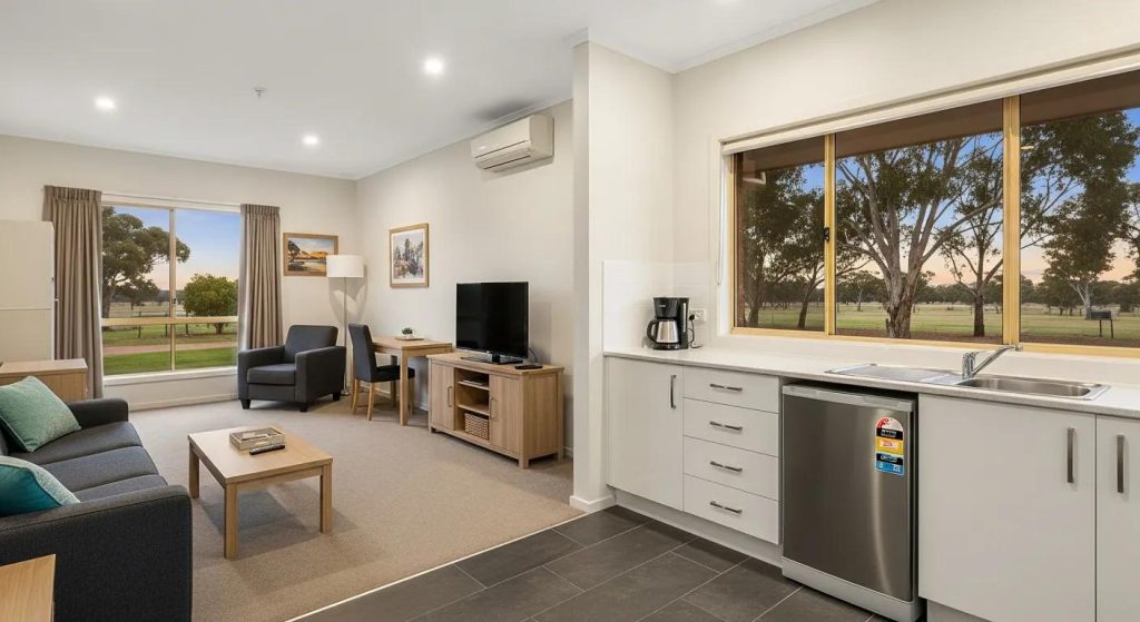 A dramatic wide-screen interior and exterior blend showcasing modern serviced accommodation in Inglewood, Victoria. A spacious, well-lit serviced suite with soft natural light, clean contemporary furnishings and a kitchenette transitions into an exterior view of a peaceful country setting. Warm evening tones, native trees and open rural landscape highlight comfort, independence and homelike convenience. No text.